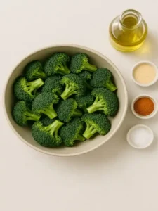 Fresh broccoli florets in a bowl next to olive oil, garlic powder, paprika, and salt on a clean kitchen countertop